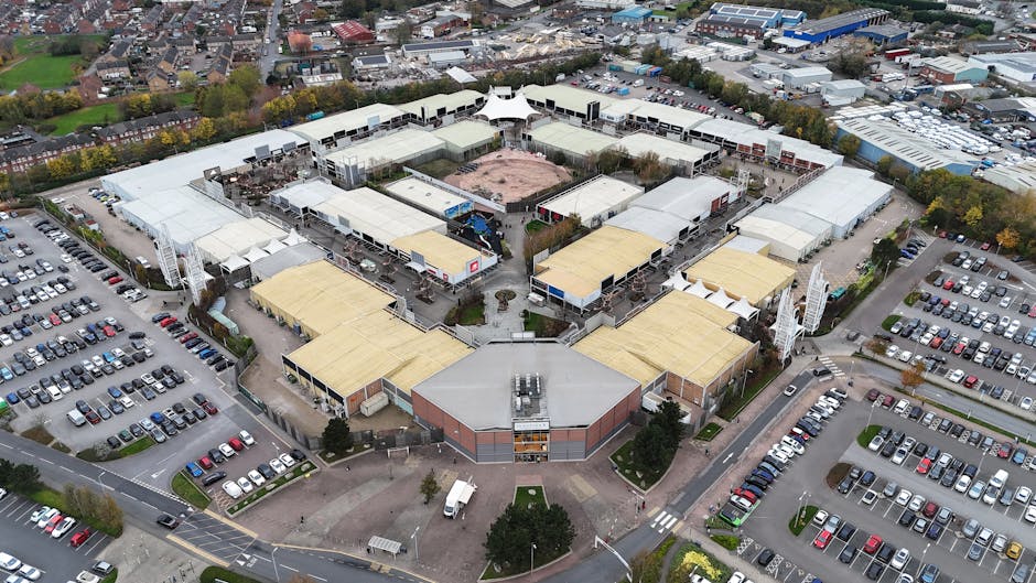 An aerial view of Brent Park retail park showcasing landscaped outdoor areas with neatly trimmed grass lawns, paved pedestrian pathways, and organized parking lots filled with cars. The shopping centr