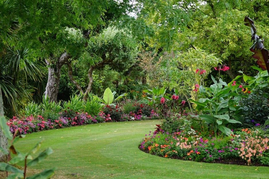 A lush garden scene featuring a well-maintained, vibrant green lawn bordered by an array of flowering plants and shrubs. On the left, a large tree with textured, weathered bark and expansive green lea